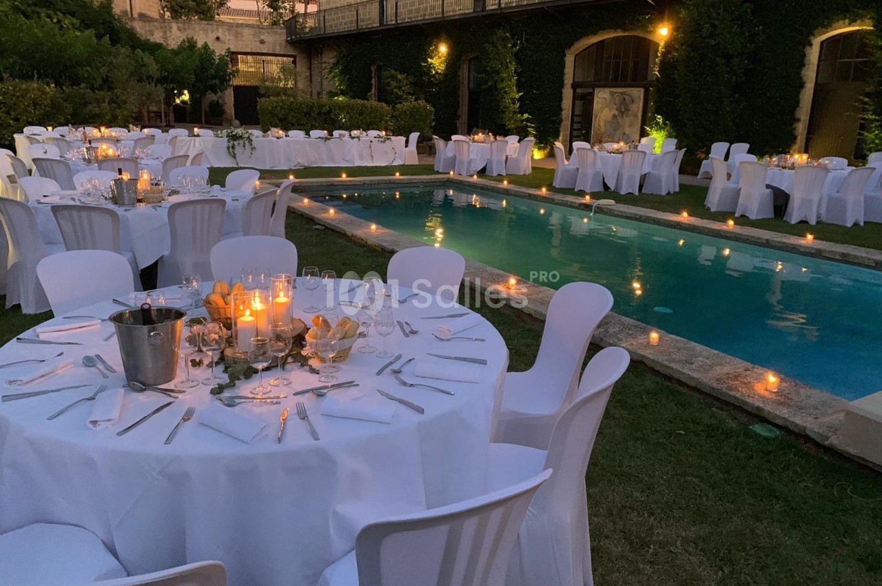 Tables dressées avec nappes blanches autour d'une piscine éclairée par des bougies dans un jardin en soirée.
