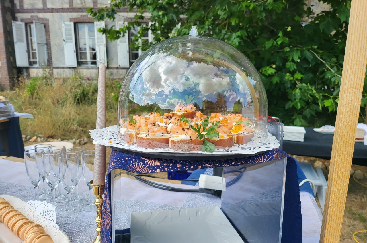 Plateau de canapés apéritifs sous une cloche en verre, disposé sur une table dressée en extérieur.