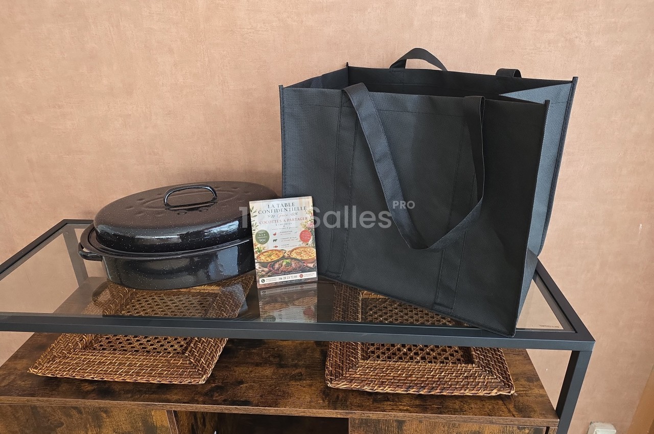 Cocotte noire avec couvercle, sac réutilisable noir et dépliant posés sur une table en verre avec sets en osier.