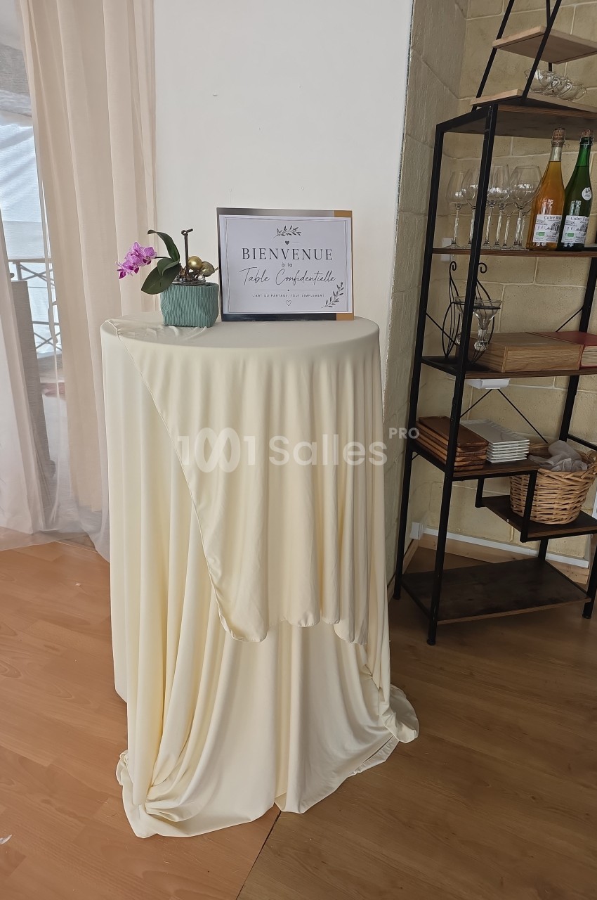 Table haute recouverte d'une nappe beige, décorée d'une orchidée et d'un cadre ’Bienvenue’, près d'une étagère.