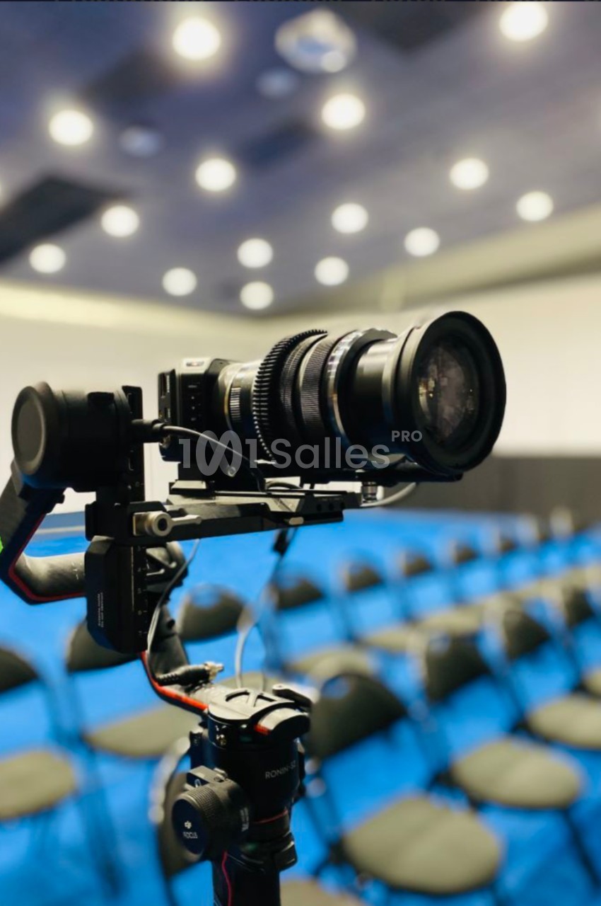 Caméra professionnelle montée sur un stabilisateur, avec des rangées de chaises floues en arrière-plan.