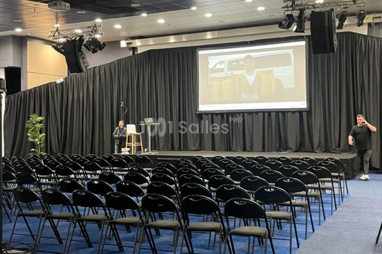 Salle de conférence avec des rangées de chaises vides, une scène équipée d'un écran géant et deux personnes présentes.