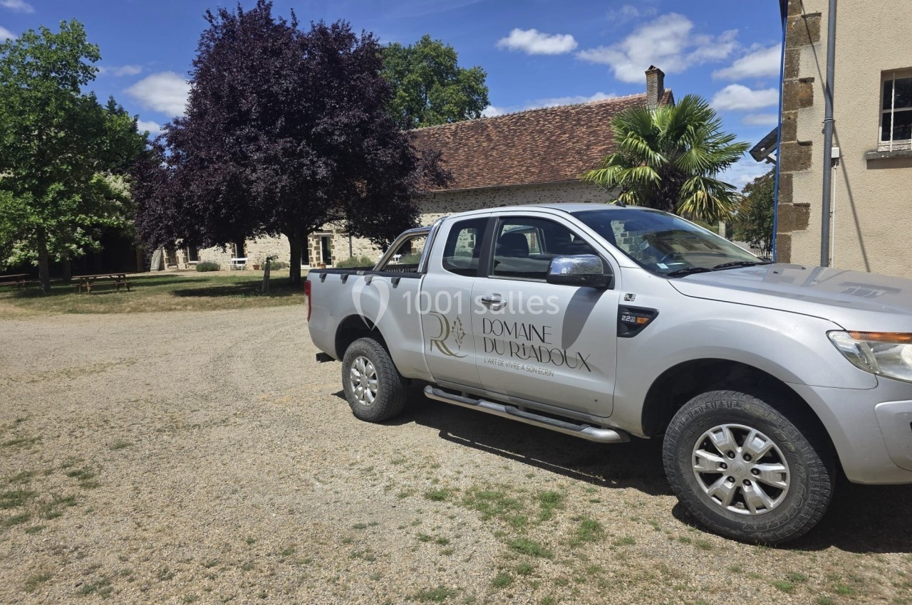 Pick-up gris stationné devant un bâtiment en pierre avec toit en tuiles, entouré d'arbres et d'un ciel dégagé.