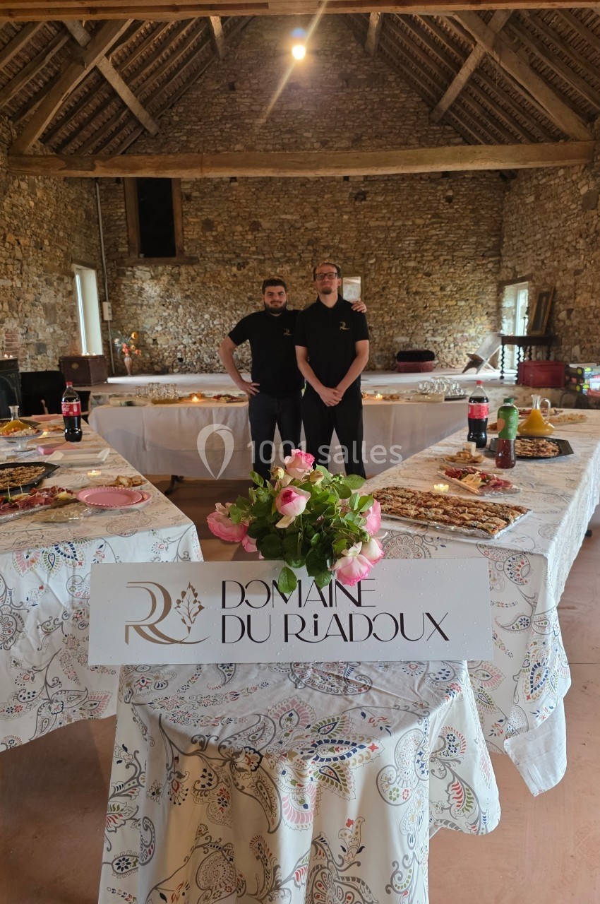 Deux personnes posent devant des tables garnies de plats dans une salle en pierre avec une enseigne ’Domaine du Riadoux’.