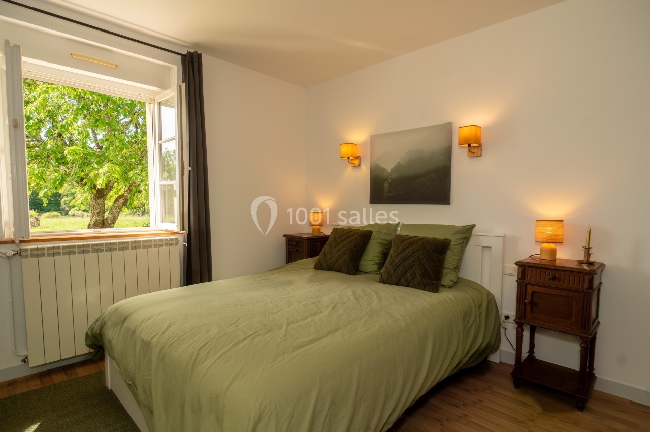 Chambre lumineuse avec lit double, draps verts, deux tables de chevet en bois et une fenêtre ouverte sur un arbre.