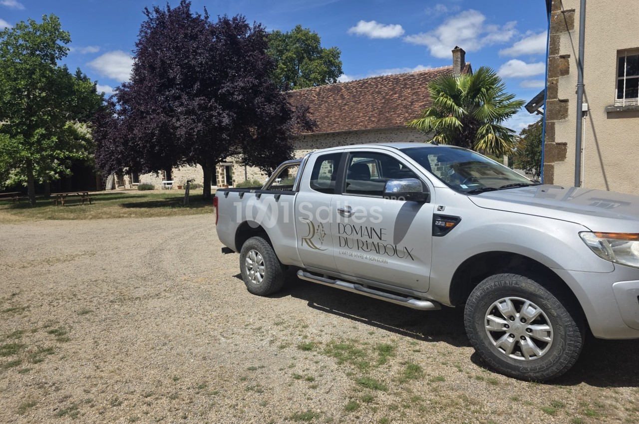 Pick-up gris stationné devant un bâtiment en pierre avec un toit en tuiles, entouré d'arbres et d'herbe.