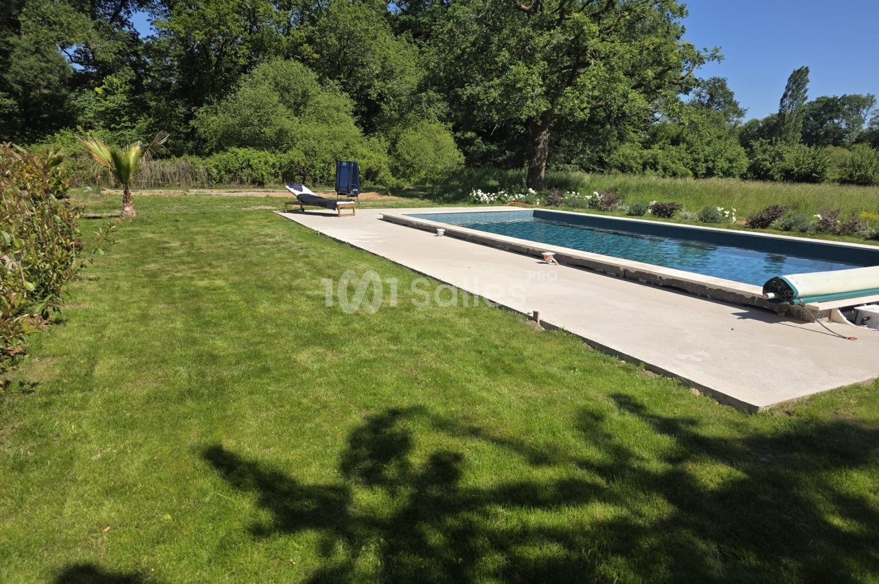 Piscine extérieure entourée de pelouse et de végétation, avec transat et parasol sous un ciel dégagé.