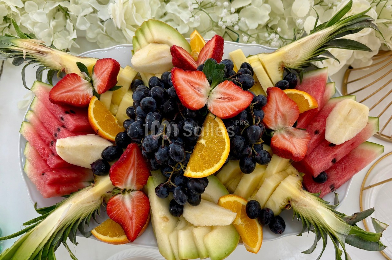 Assortiment de fruits frais comprenant fraises, raisins, pastèque, ananas, bananes et tranches d'orange sur un plateau.