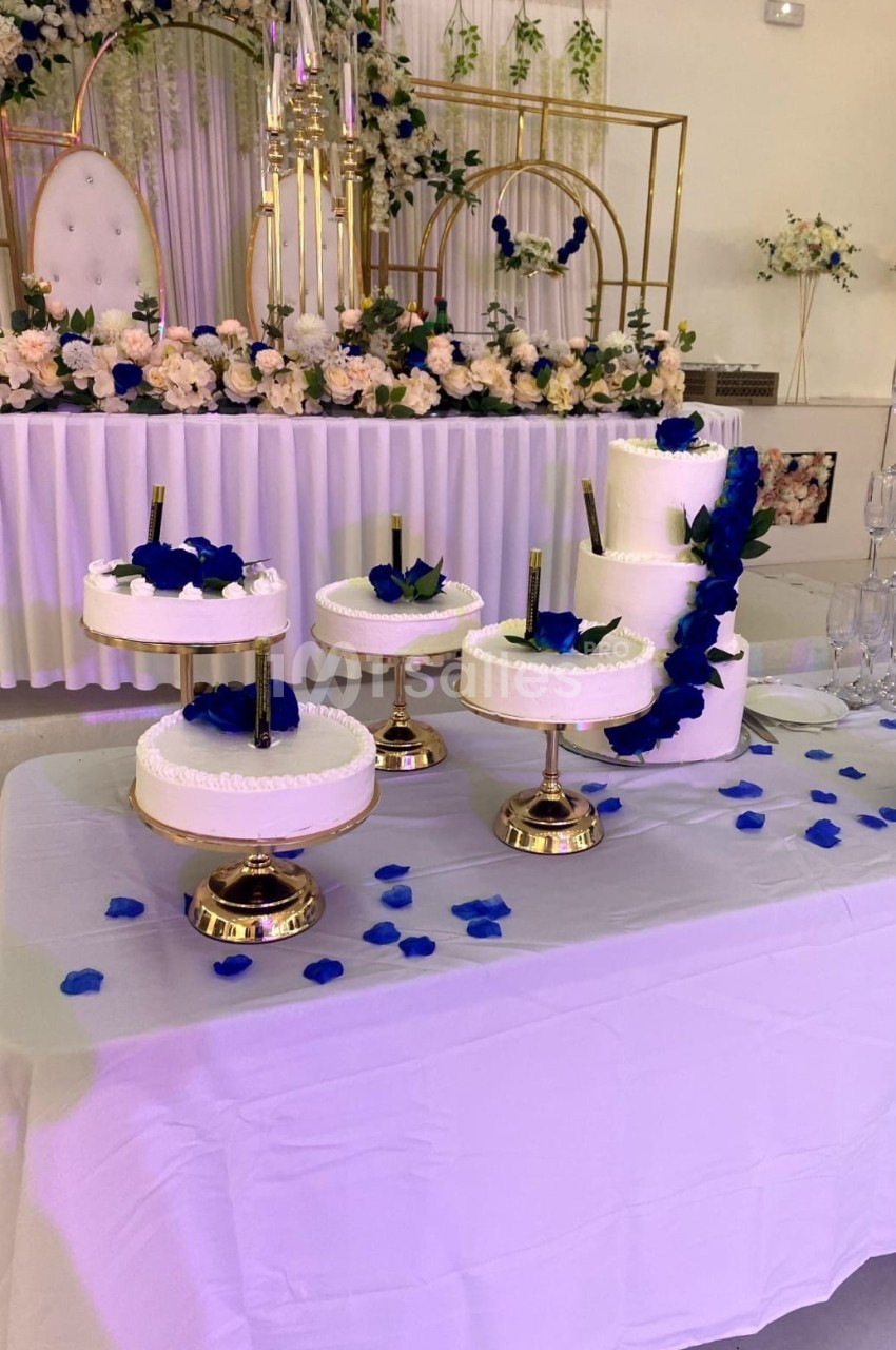 Table décorée avec plusieurs gâteaux blancs sur des présentoirs dorés, ornés de pétales et fleurs bleues.