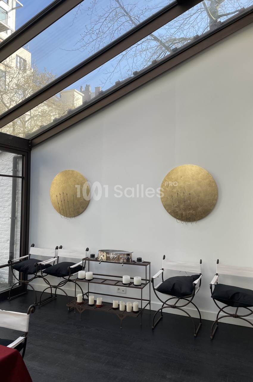 Salle lumineuse avec verrière, murs blancs décorés de disques dorés, chaises design et étagère avec bougies.