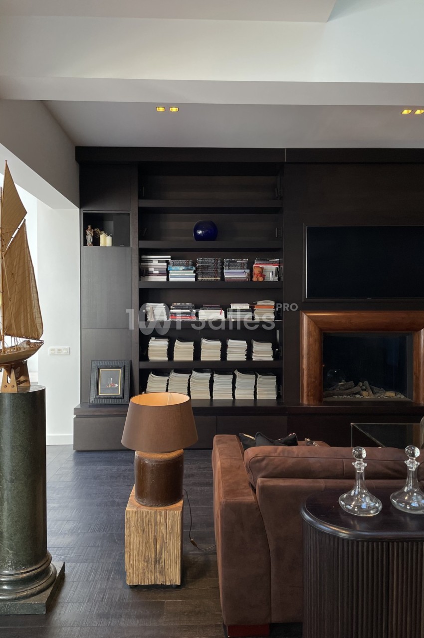 Salon moderne avec bibliothèque remplie de livres, canapé marron, lampe sur table et maquette de bateau sur un socle.