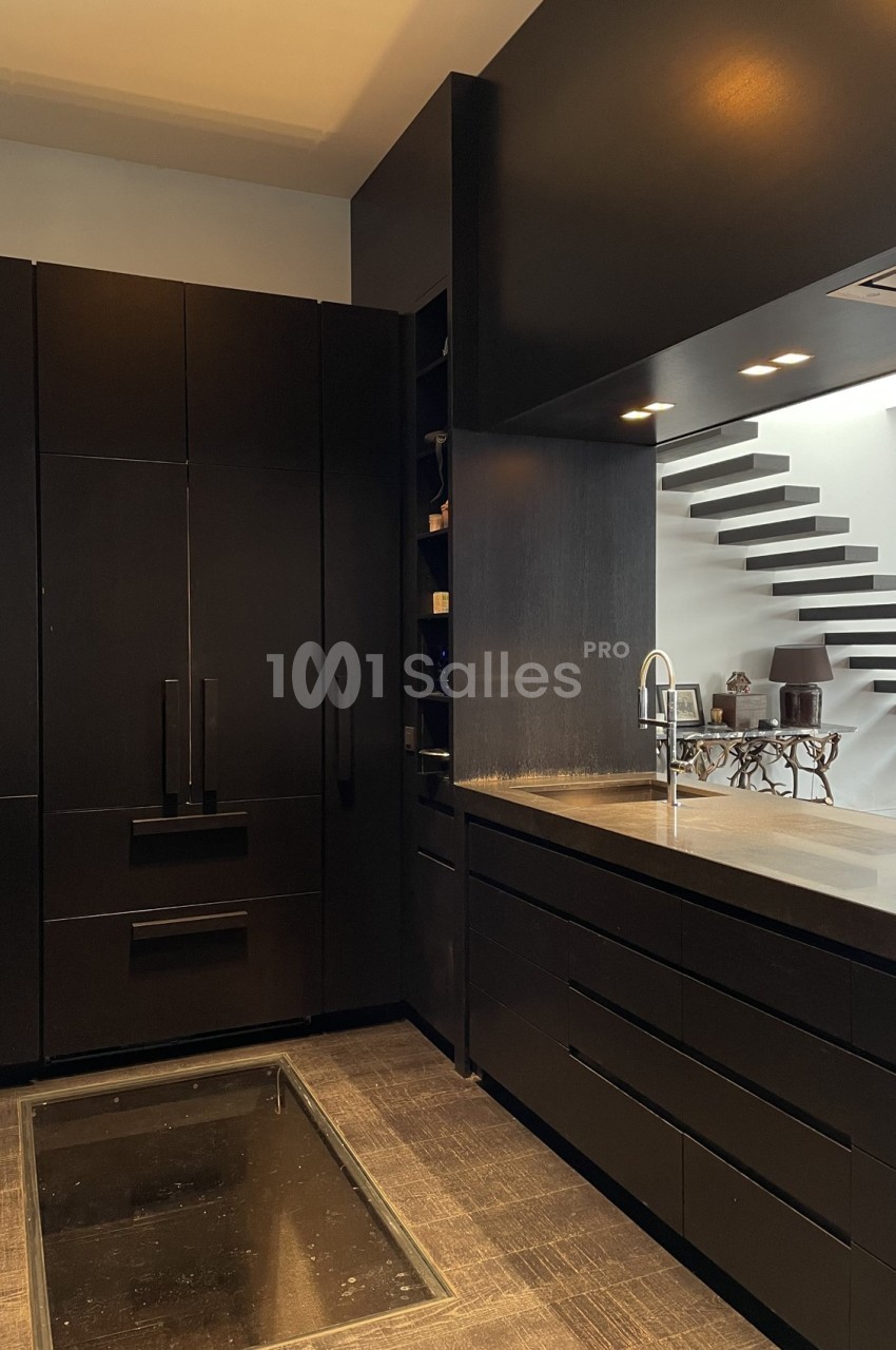 Cuisine moderne avec meubles noirs, plan de travail sombre, évier en inox et escalier minimaliste en arrière-plan.