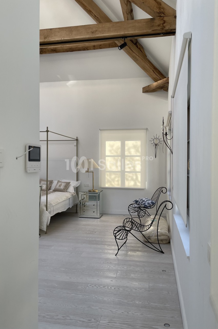 Chambre lumineuse avec lit à baldaquin, fauteuil en métal, poutres apparentes et fenêtre laissant entrer la lumière.