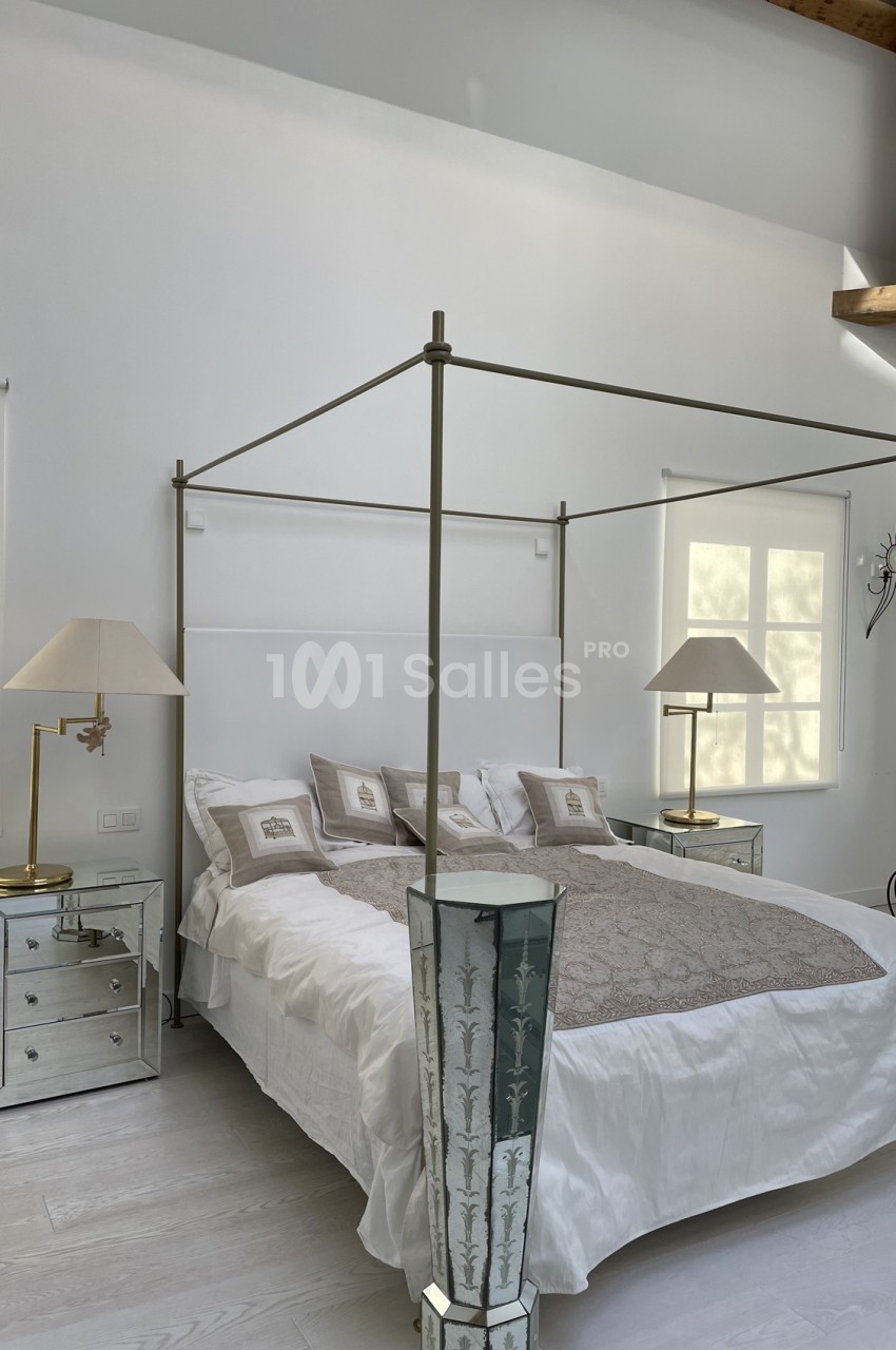 Lit à baldaquin moderne avec draps blancs, tables de chevet en miroir et lampes, dans une chambre lumineuse.