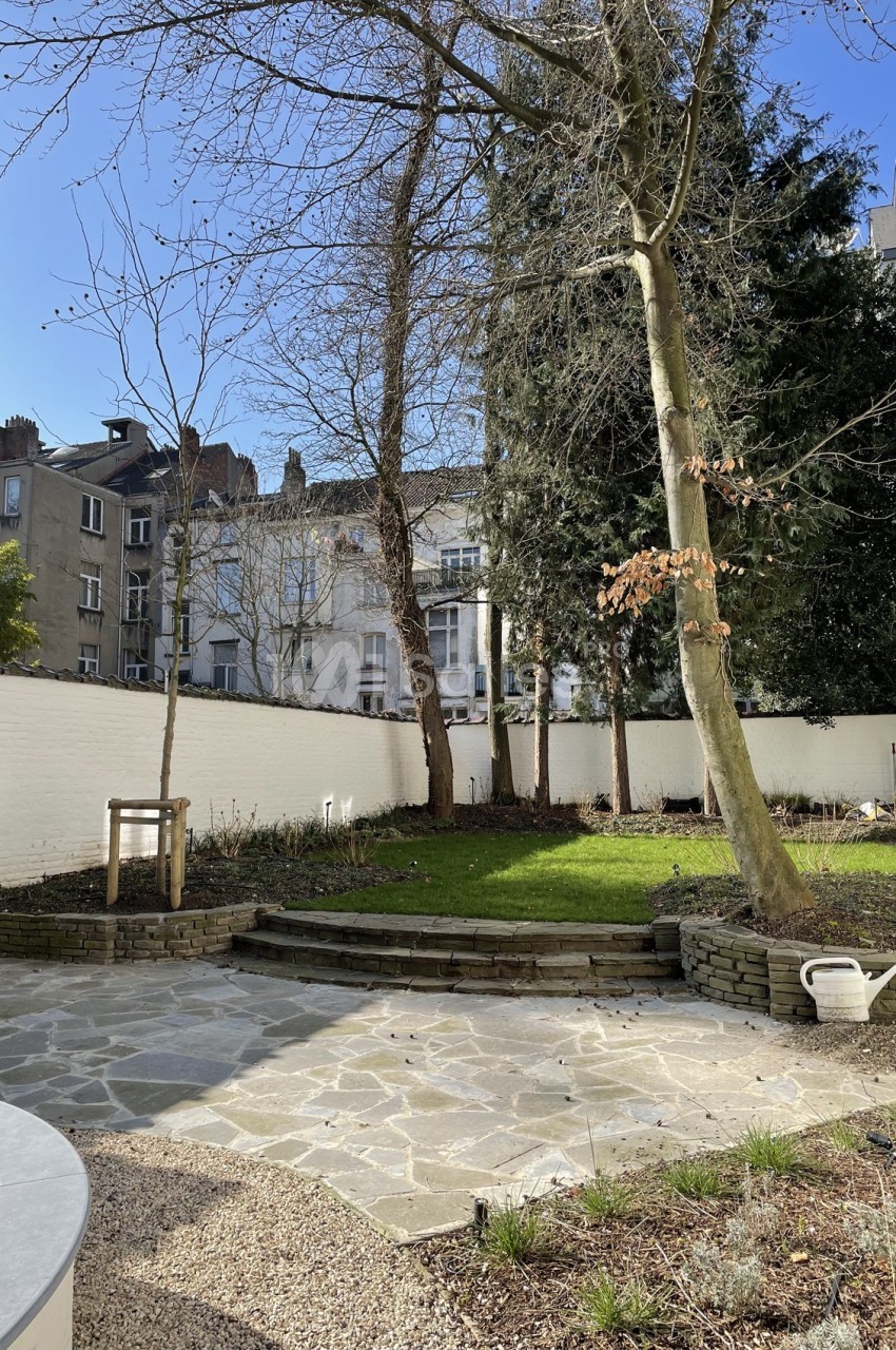 Jardin urbain avec pelouse, arbres, muret en pierre et sol pavé, entouré de murs blancs et d'immeubles en arrière-plan.