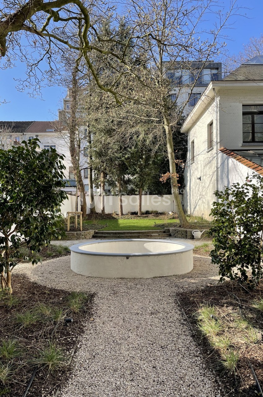 Petit jardin avec un chemin de gravier menant à une fontaine circulaire entourée de végétation et d'arbres.
