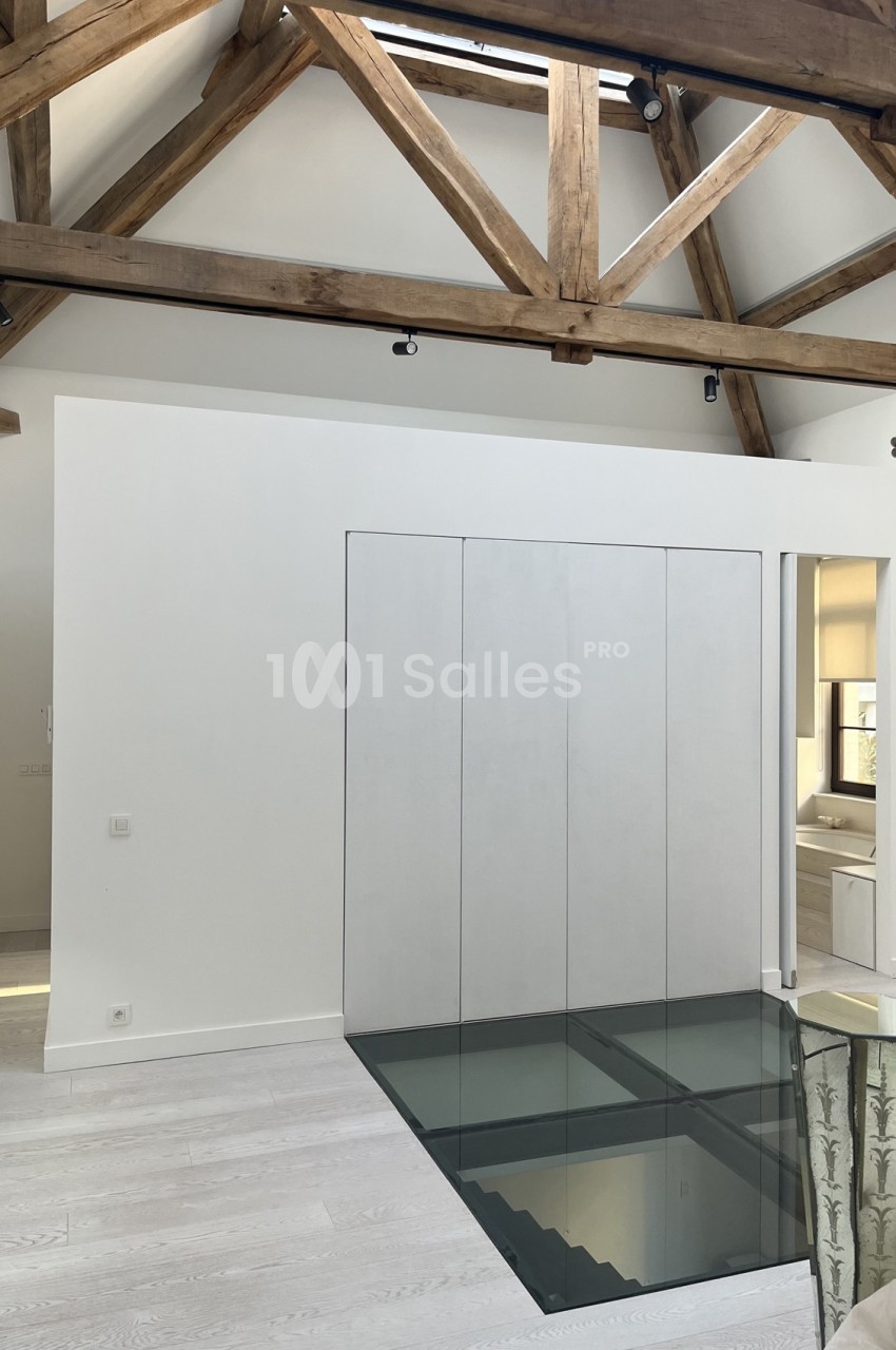 Plancher en verre transparent dans un intérieur moderne avec poutres apparentes en bois et murs blancs.