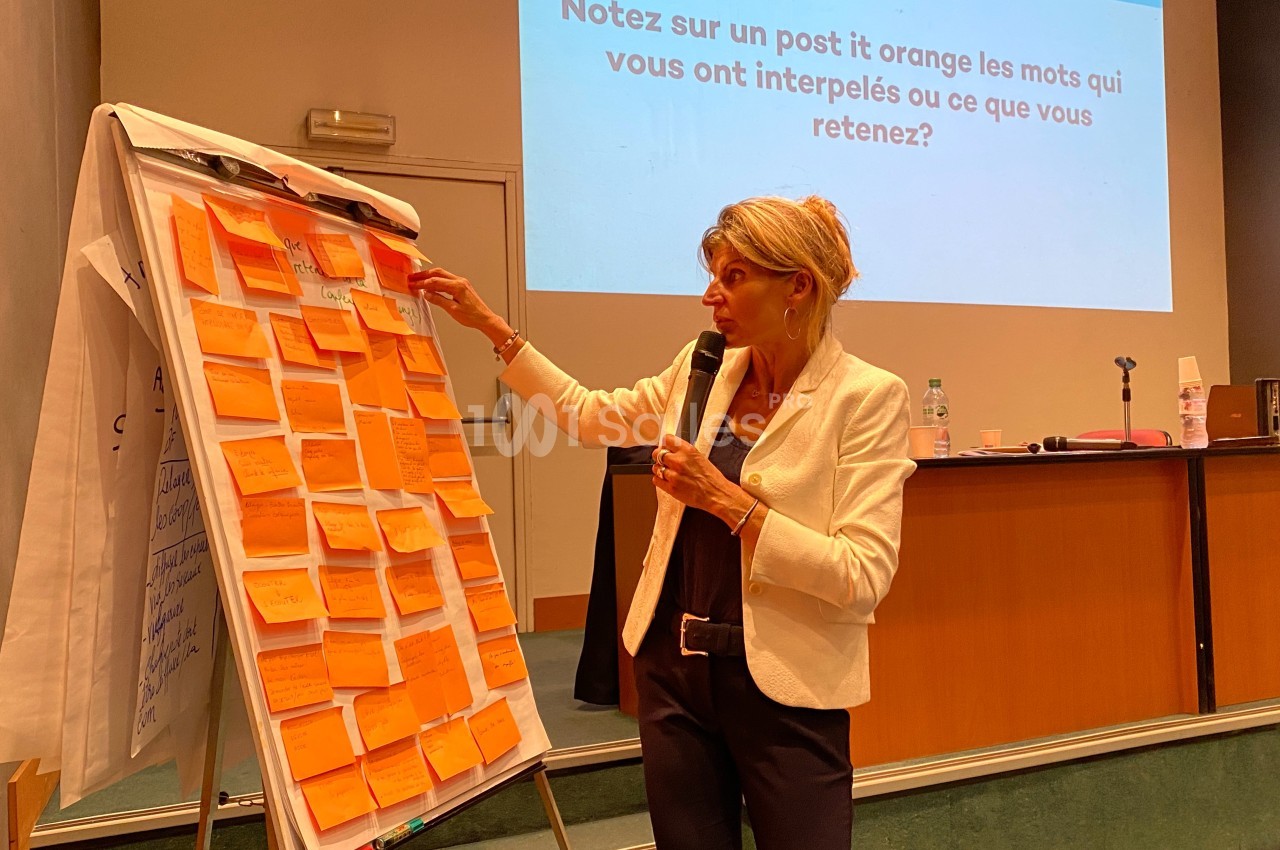 Une femme présente devant un paperboard rempli de post-it orange dans une salle de conférence.