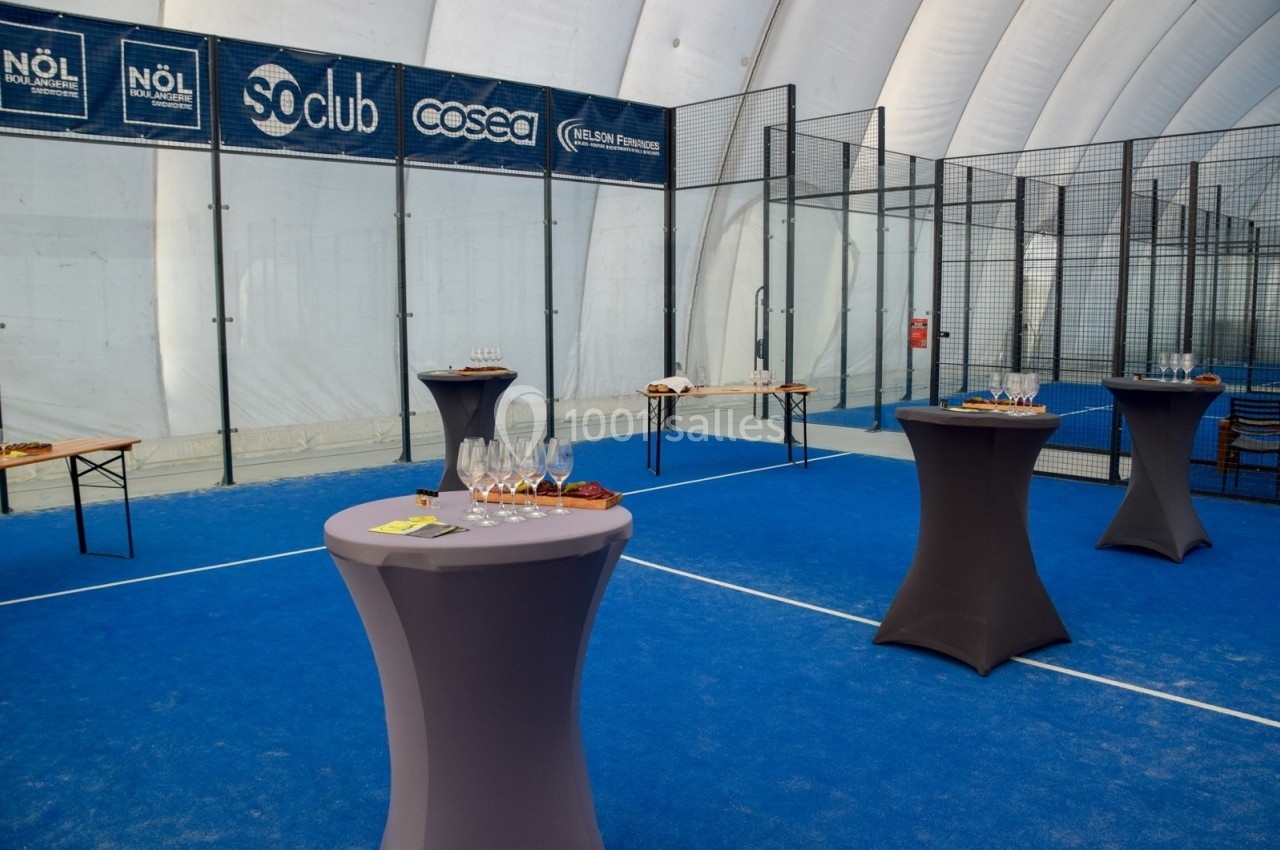 Tables hautes avec verres et amuse-bouches disposées sur un terrain de padel couvert pour un événement.