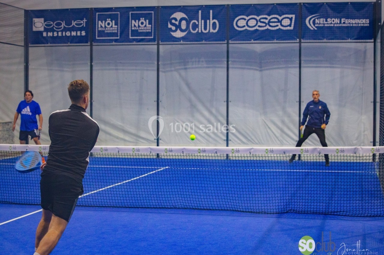 Deux joueurs de padel en plein échange sur un terrain intérieur, un troisième joueur observe en arrière-plan.