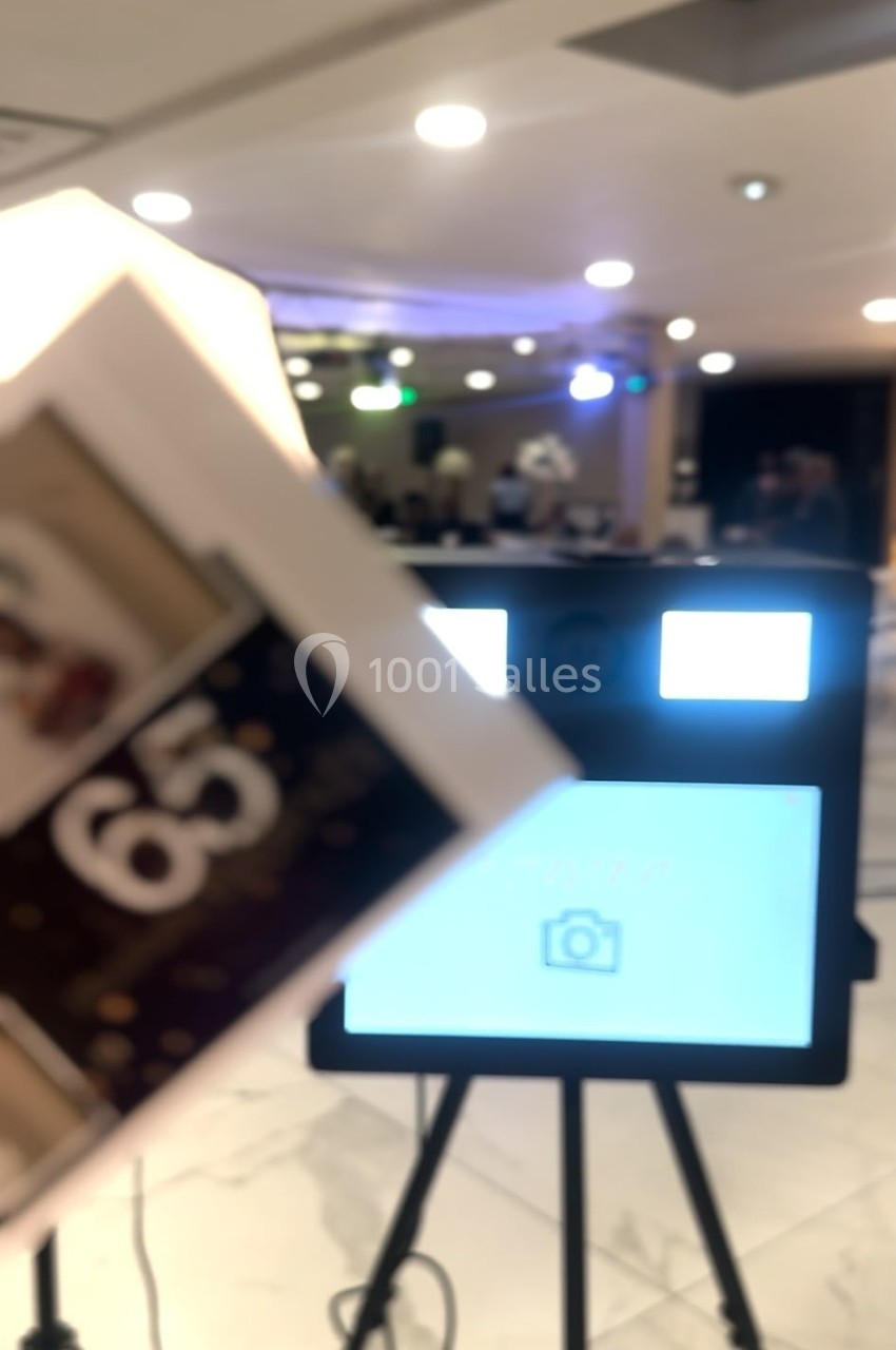 Photo floue d'une borne photo sur trépied avec un écran lumineux, en arrière-plan une salle éclairée.