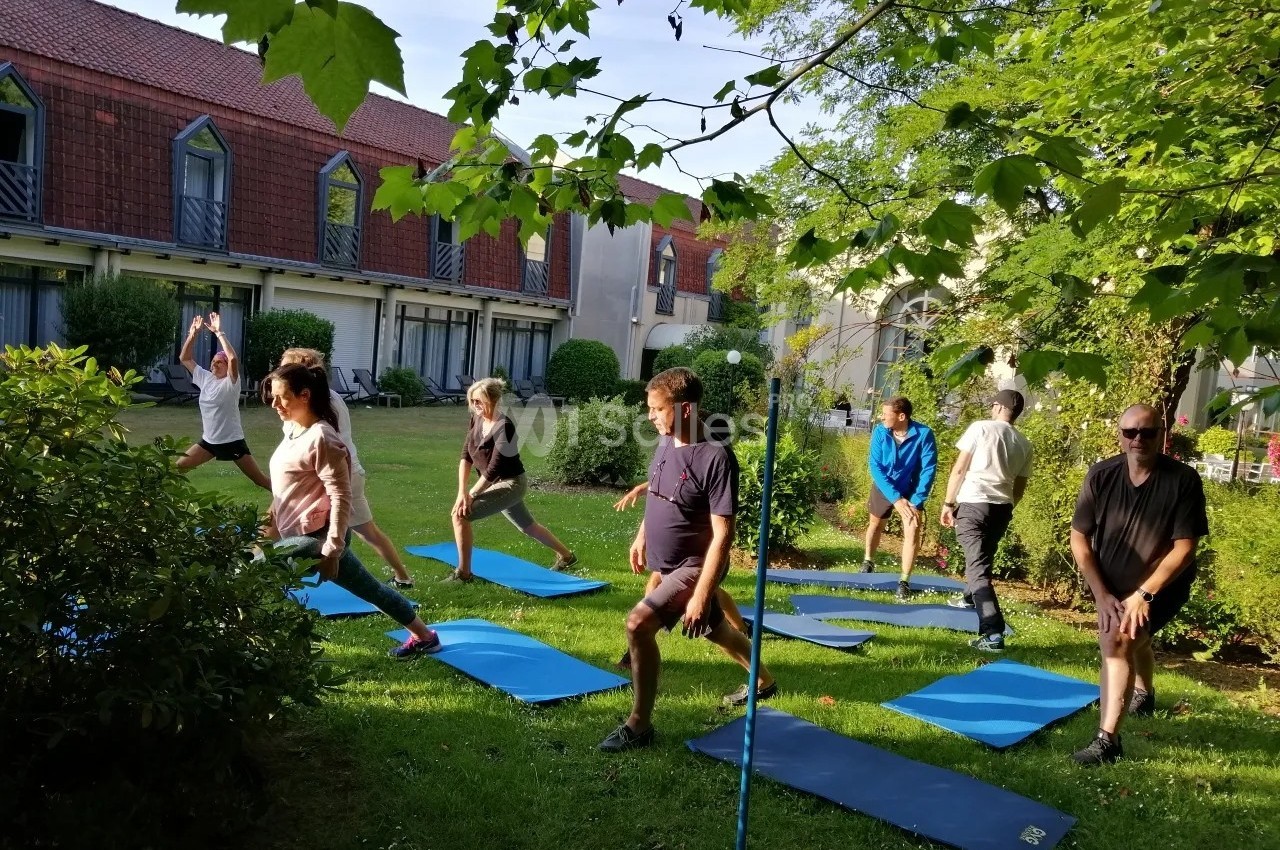 Personnes pratiquant des exercices de yoga en plein air sur des tapis, dans un jardin entouré de verdure et de bâtiments.