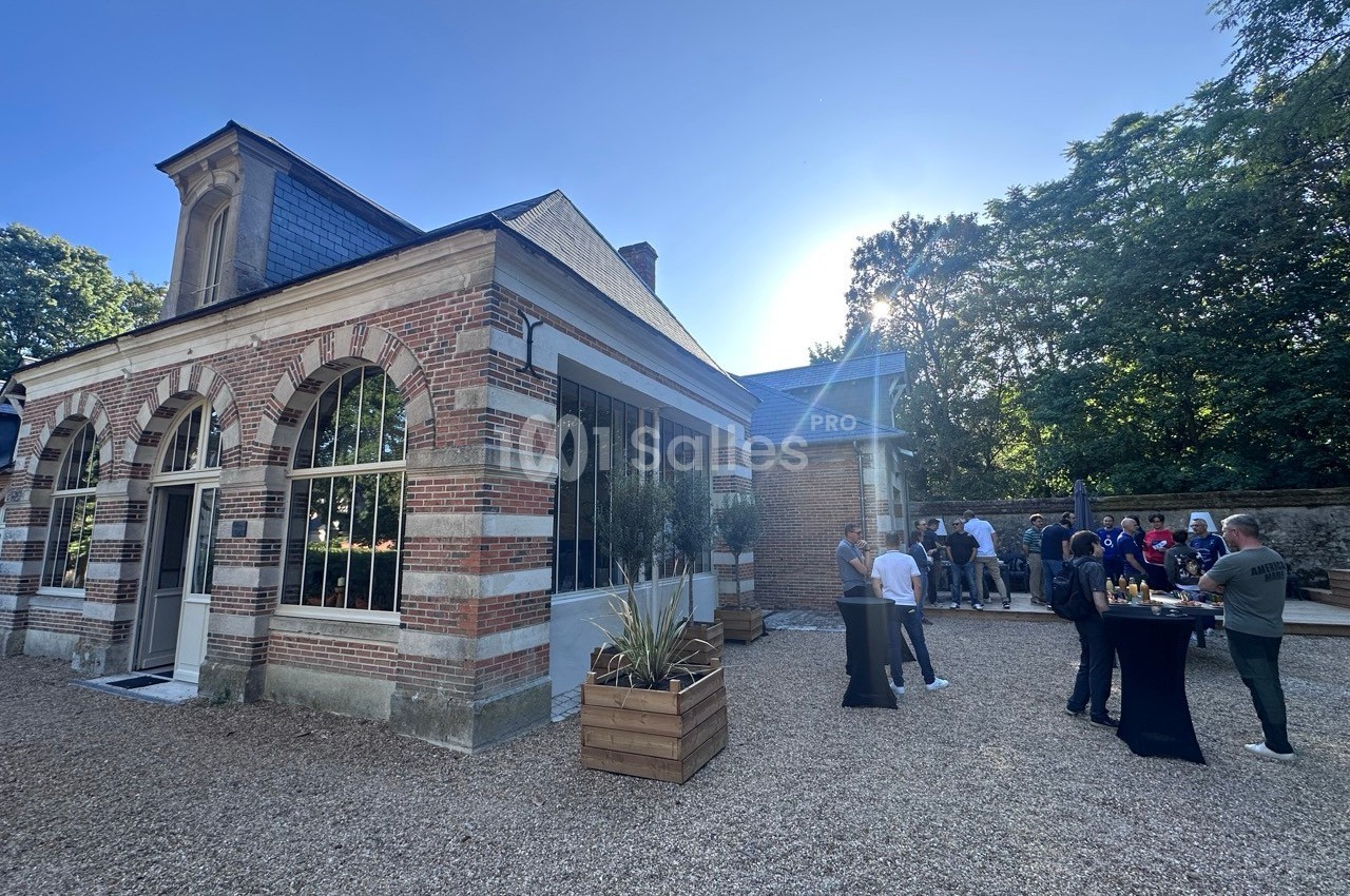 Bâtiment en briques avec grandes fenêtres, terrasse en gravier, personnes discutant autour de tables hautes.
