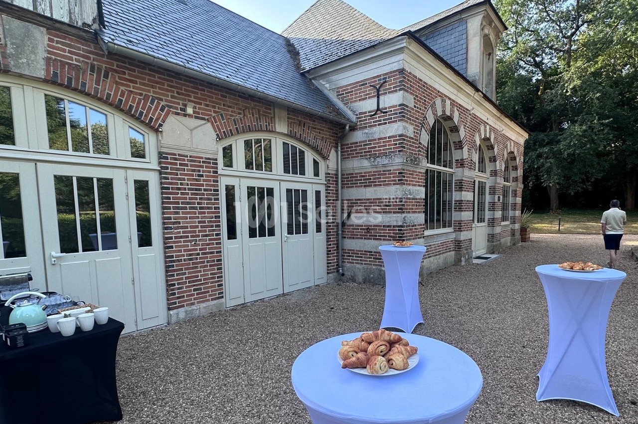 Table dressée avec viennoiseries et boissons devant un bâtiment en briques et grandes fenêtres, dans une cour gravillonnée.