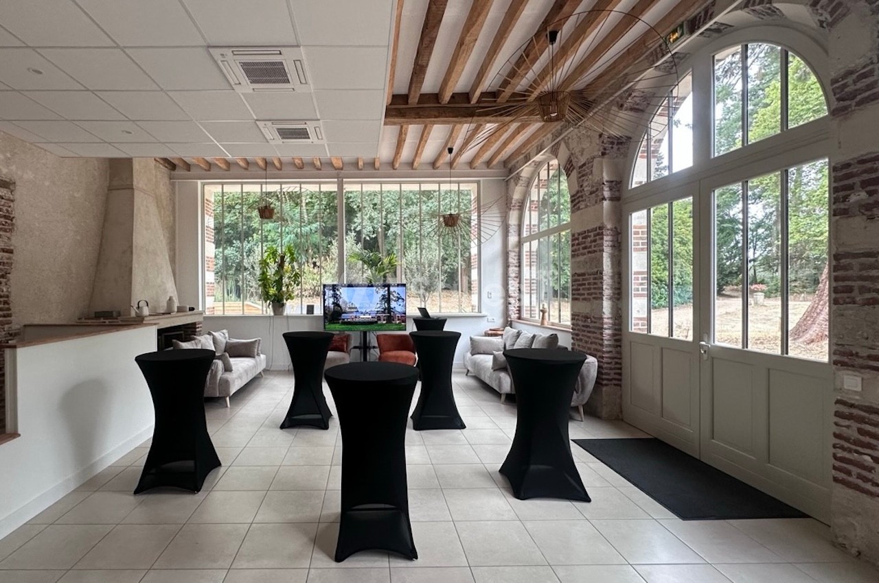 Salle lumineuse avec grandes baies vitrées, tables hautes noires, canapés et écran affichant une image.