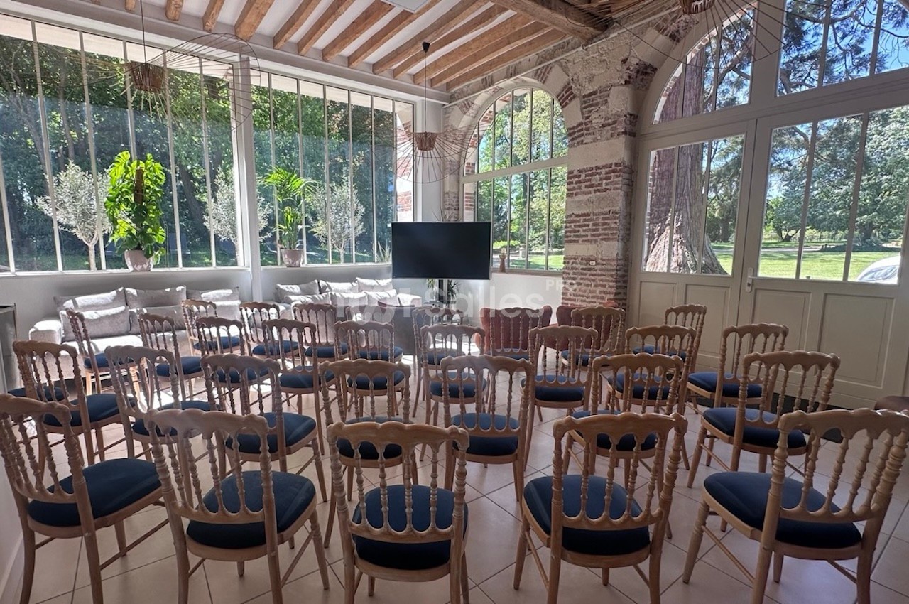 Salle lumineuse avec chaises en bois disposées face à un écran, grandes fenêtres donnant sur un jardin verdoyant.