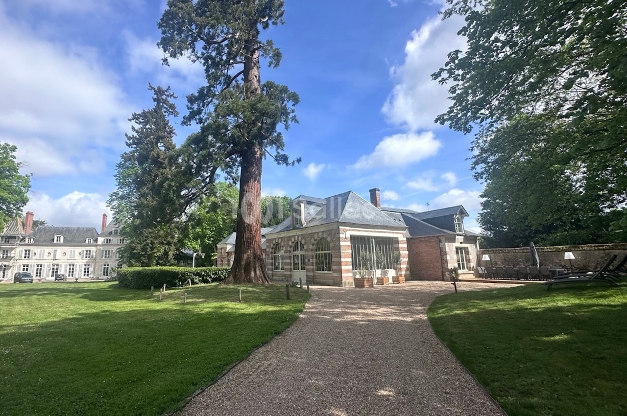 Allée en gravier menant à un bâtiment en pierre entouré de pelouses et d'arbres sous un ciel partiellement nuageux.