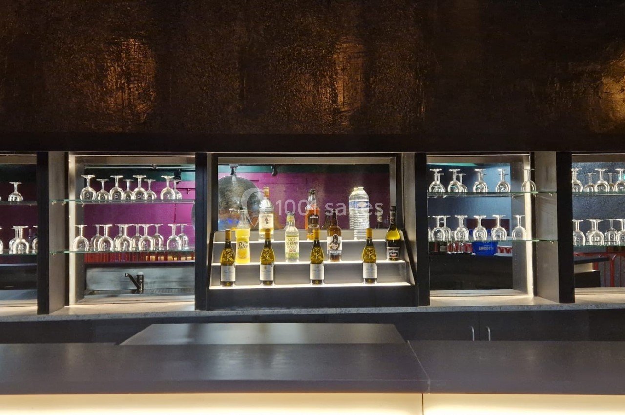 Bar avec étagères illuminées contenant des verres et des bouteilles, sur fond sombre avec éclairage tamisé.