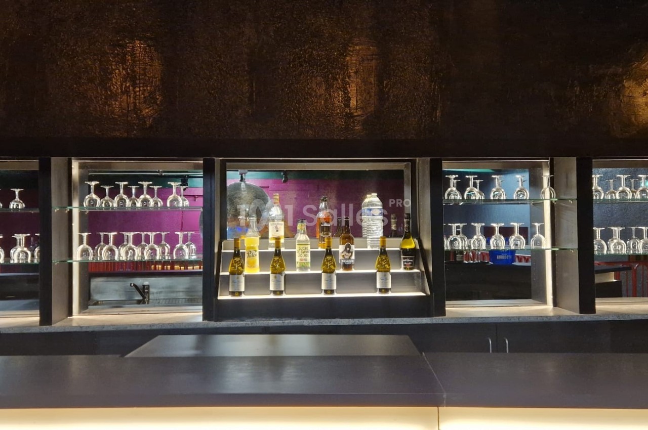 Bar moderne avec étagères en verre illuminées, exposant des verres et des bouteilles d'alcool devant un mur sombre.