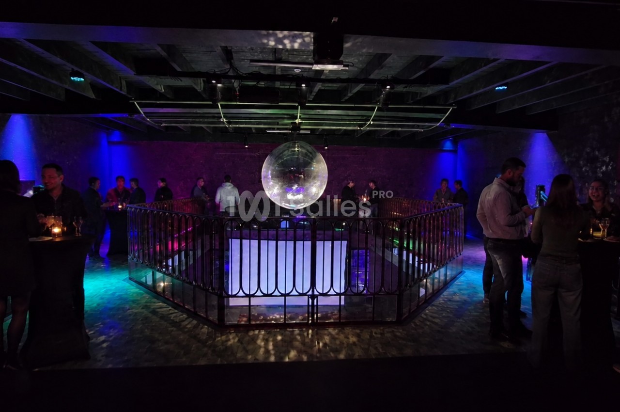 Salle sombre avec une piste de danse centrale, éclairée par une boule à facettes et des lumières colorées, entourée de…