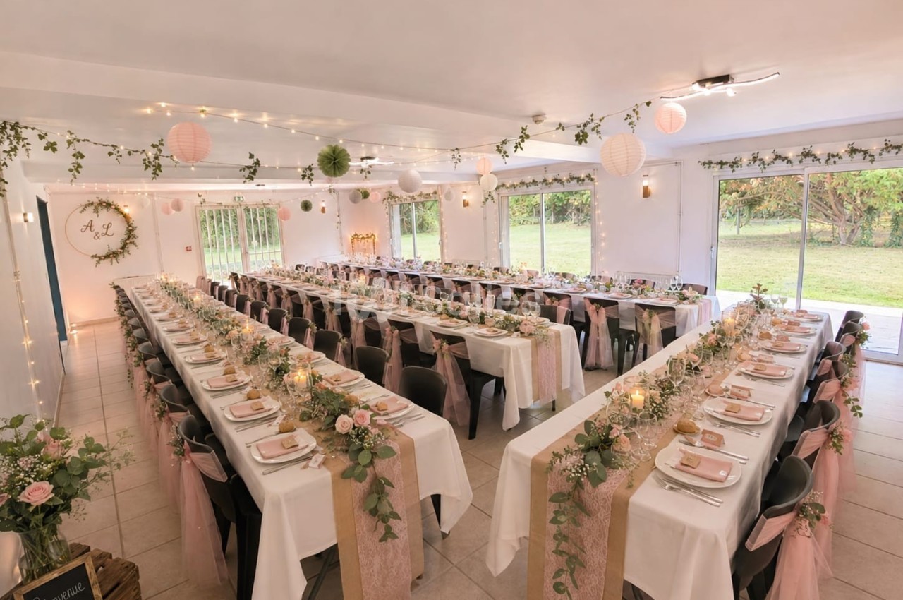 Salle de réception décorée pour un mariage, avec longues tables ornées de fleurs, nappes blanches et guirlandes lumineuses.