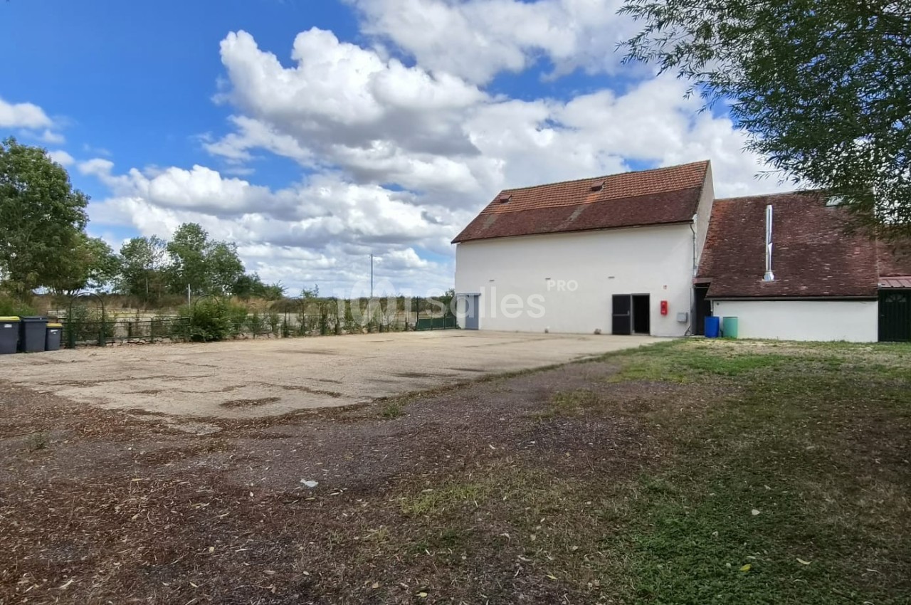 Cour extérieure avec sol en gravier, bâtiment blanc à toit rouge, arbres et poubelles en arrière-plan sous un ciel nuageux.