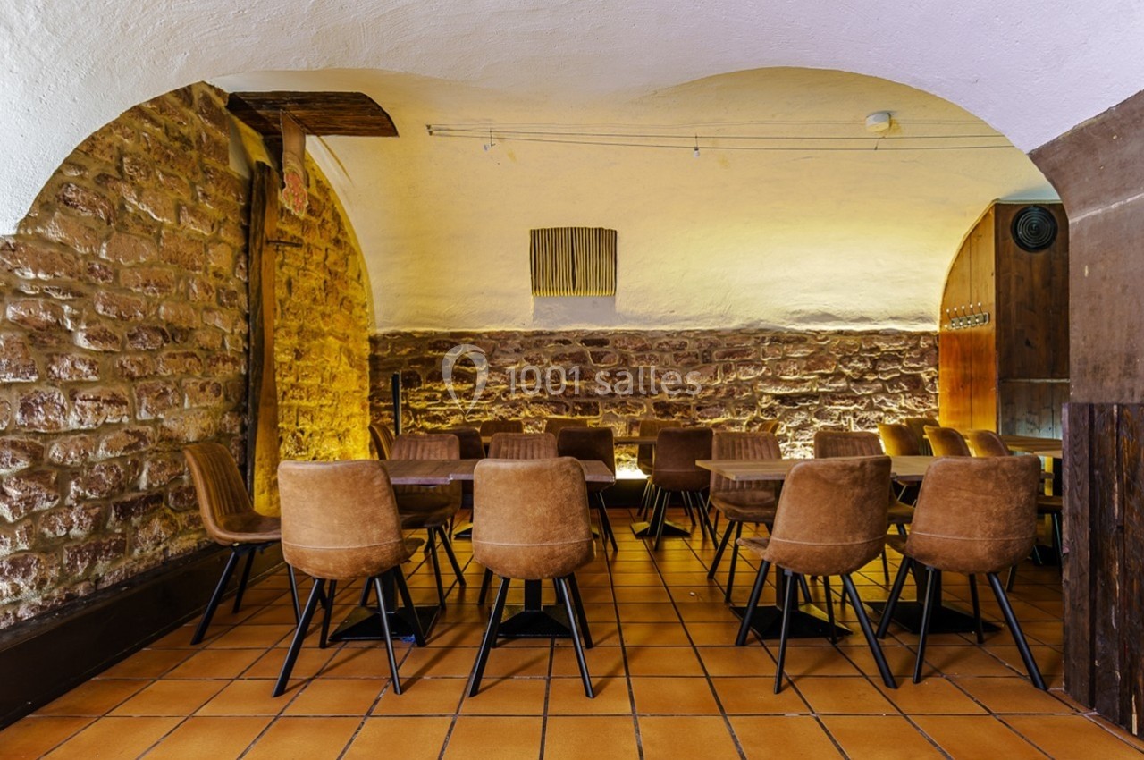 Salle voûtée en pierre avec des tables et des chaises en cuir marron disposées sur un sol en carrelage.