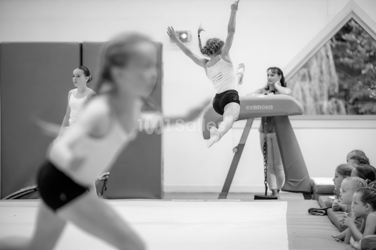 Une gymnaste effectue un saut acrobatique sur un trampoline, observée par des enfants assis au sol.