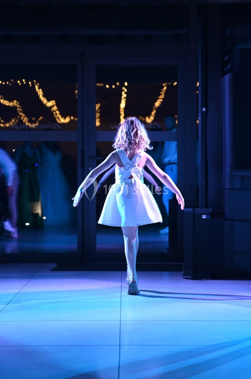 Une fillette en robe blanche danse sur une scène éclairée, avec des lumières décoratives en arrière-plan.
