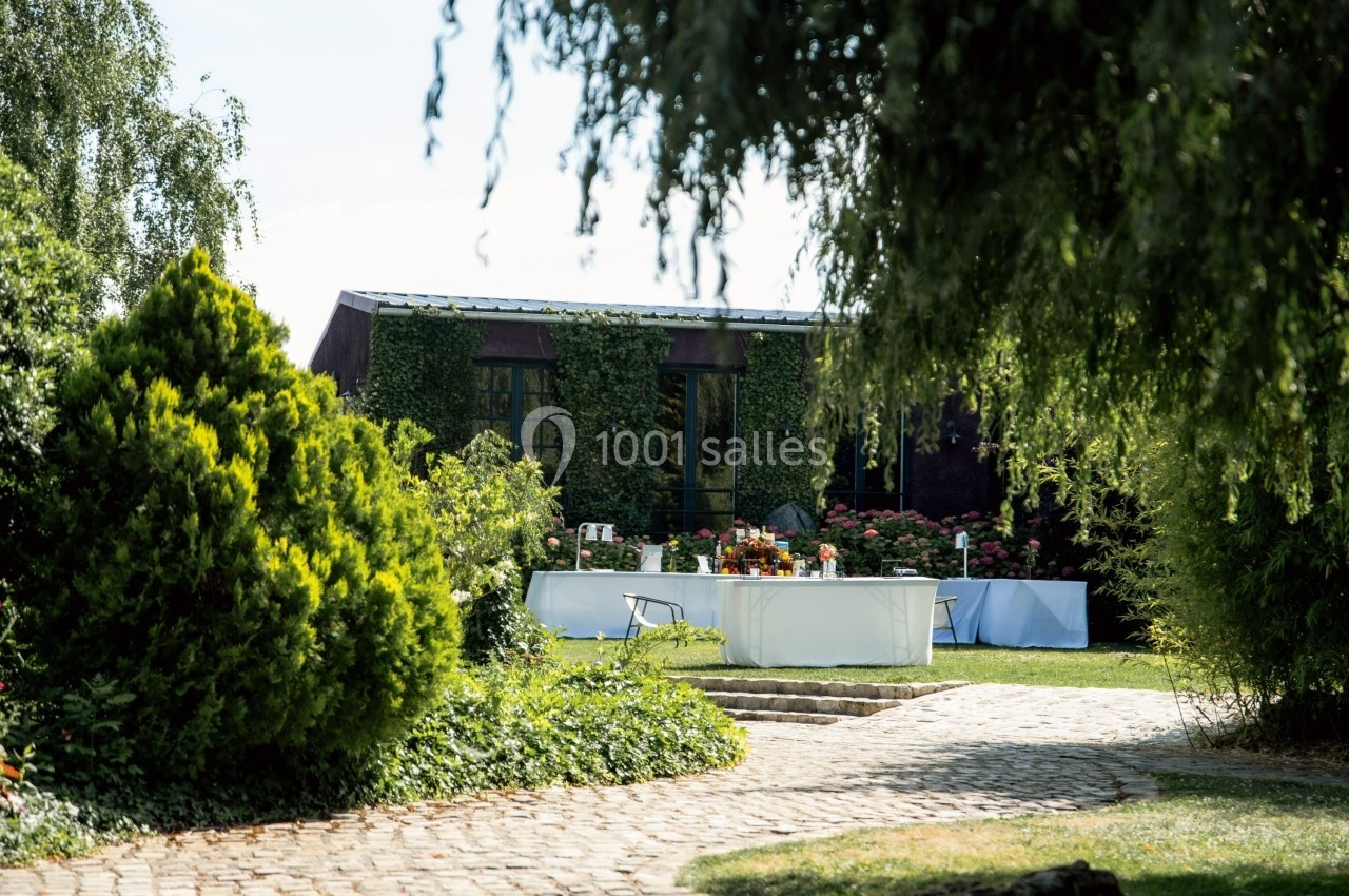 Table dressée en extérieur dans un jardin verdoyant, entourée de buissons et d'arbres sous un ciel dégagé.