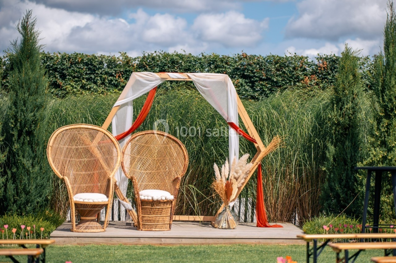 Décor extérieur avec deux fauteuils en osier sous une arche ornée de tissus et de pampas, entouré de verdure.