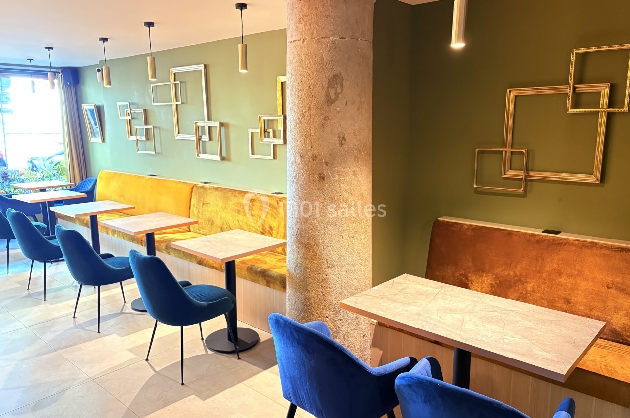 Salle de restaurant moderne avec tables en bois, chaises bleues, banquettes jaunes et murs décorés de cadres dorés.