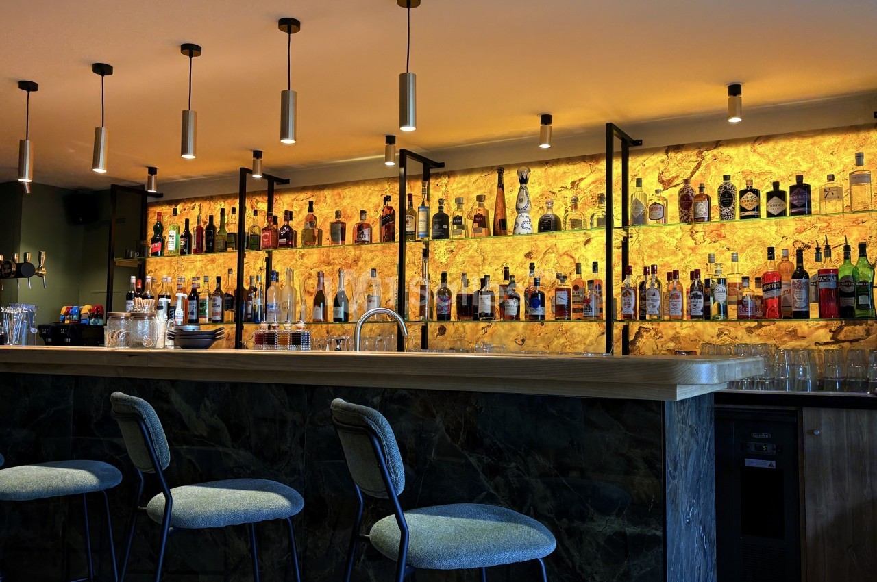 Bar moderne avec comptoir en bois, tabourets rembourrés et étagères illuminées présentant des bouteilles d'alcool.