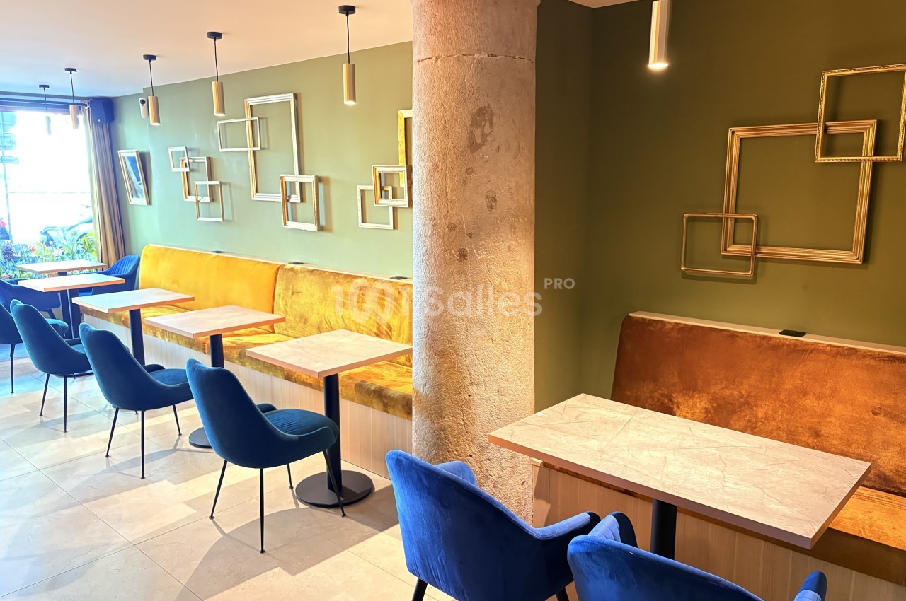 Salle de restaurant moderne avec murs verts, banquettes orange, chaises bleues et tables en bois clair.