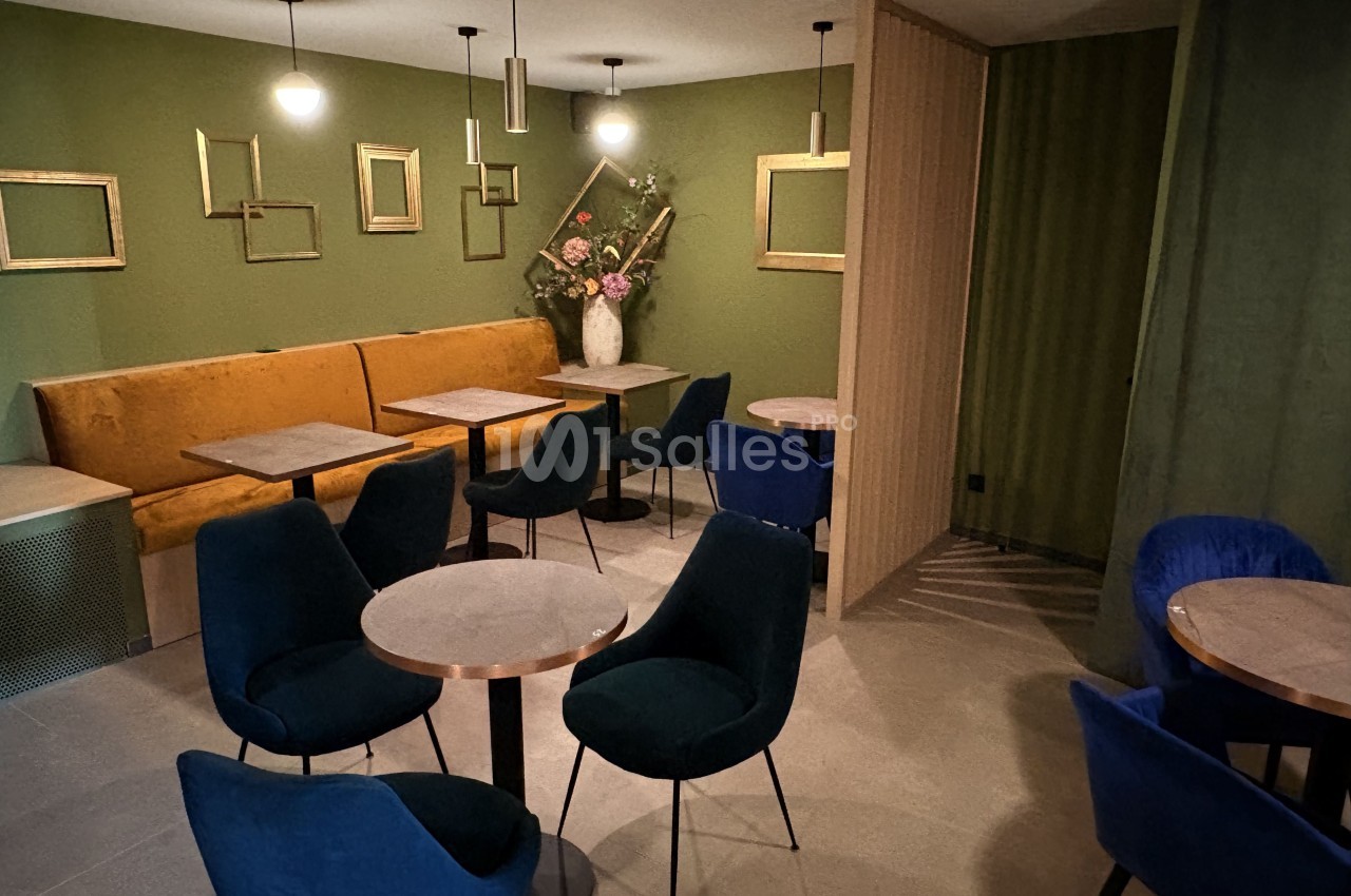 Salle de restaurant moderne avec murs verts, banquette jaune, chaises bleues et tables rondes en bois clair.