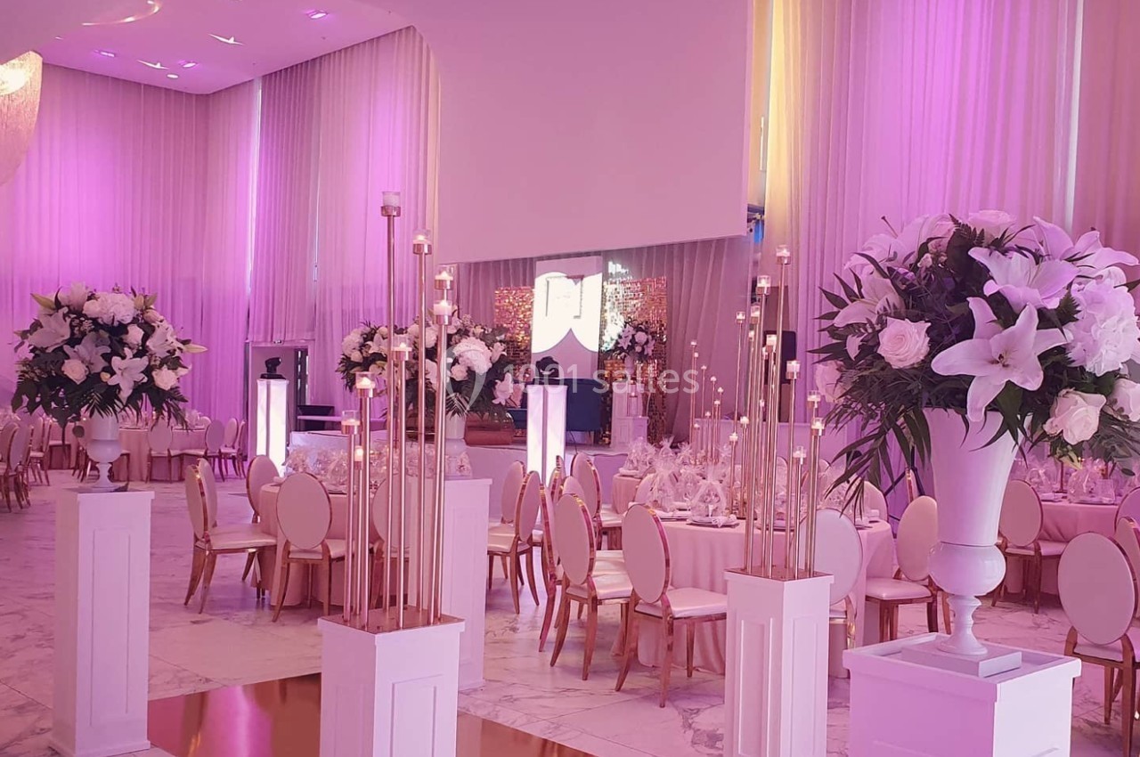 Salle de réception élégante décorée de fleurs blanches, chandeliers dorés et éclairages roses.