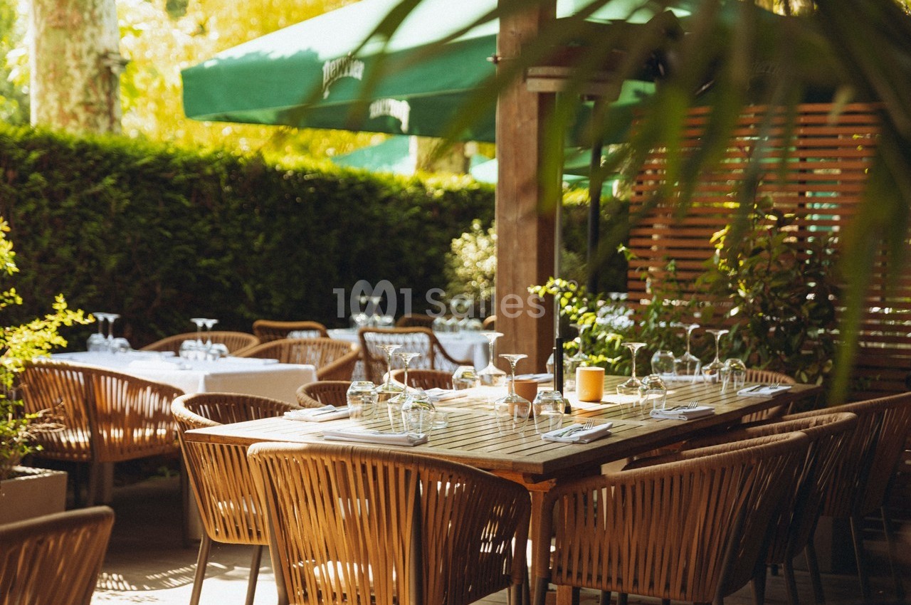 Terrasse d'un restaurant avec des tables en bois dressées, entourée de végétation et protégée par des parasols verts.