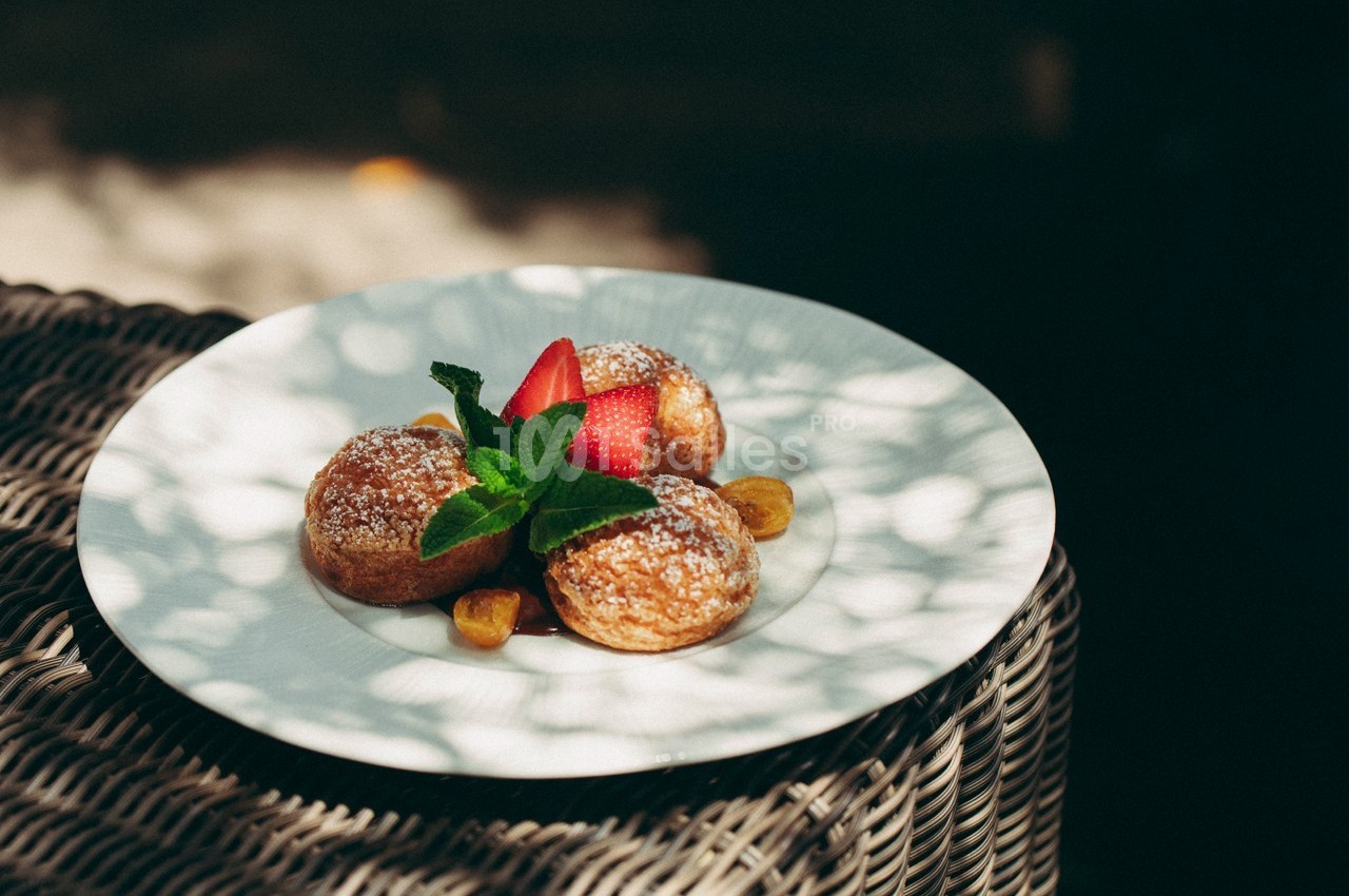 Assiette de beignets saupoudrés de sucre glace, garnis de fraises, feuilles de menthe et accompagnés de morceaux de fruits.
