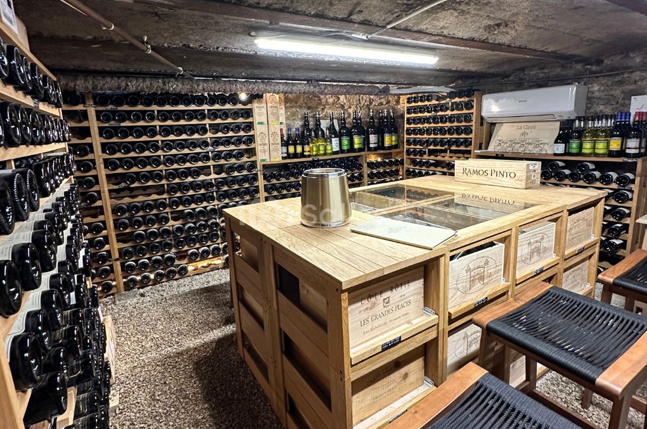 Cave à vin avec étagères remplies de bouteilles, table en bois au centre et tabourets en premier plan.