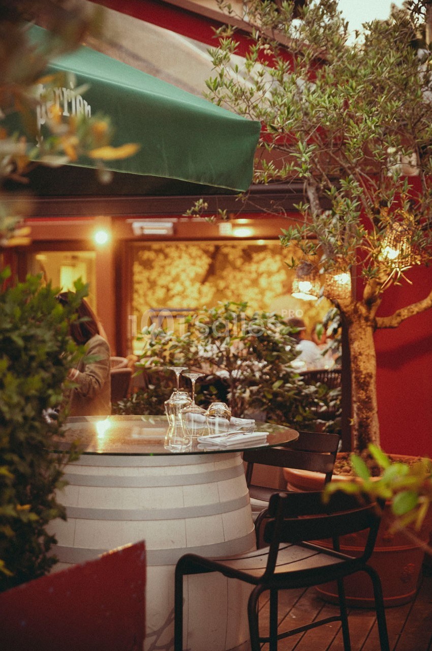 Terrasse de restaurant avec table ronde en verre sur un tonneau, chaises noires et végétation environnante.